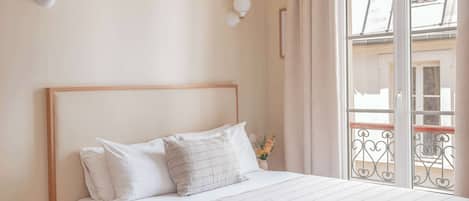 A bedroom with a bed, bedside table, wall-mounted light fixtures, and a window with a view of buildings.