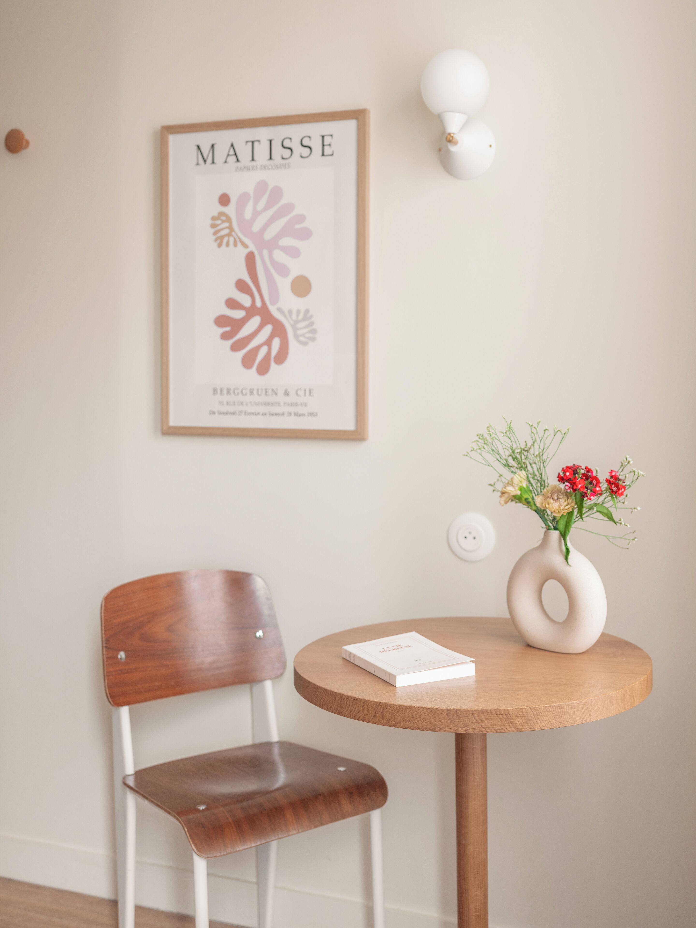 A room with a wooden chair, a small round table with a vase of flowers, and a framed artwork on the wall.
