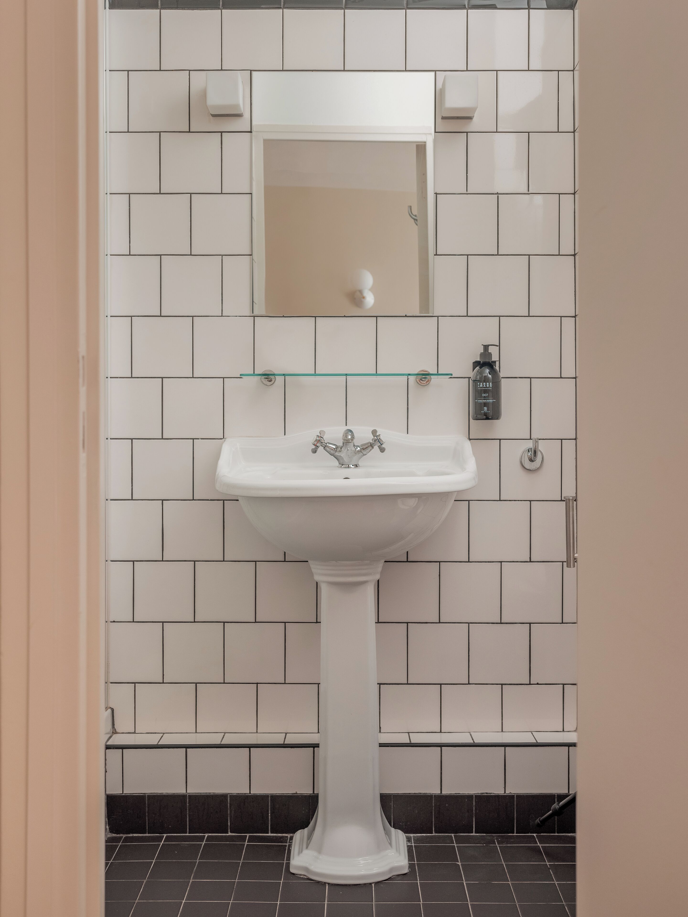 Un baño con un lavamanos pedestal blanco, un espejo encima y un dispensador de jabón en la pared.