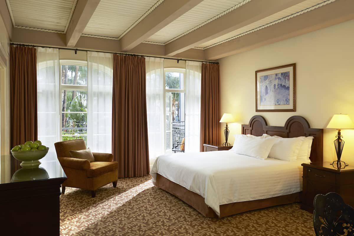 A hotel room with a large bed, two bedside lamps, a chair, a desk with a bowl of fruit, and a window with curtains.