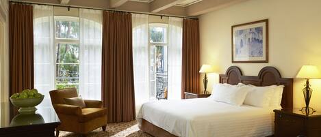 A hotel room with a large bed, two bedside lamps, a chair, a desk with a bowl of fruit, and a window with curtains.