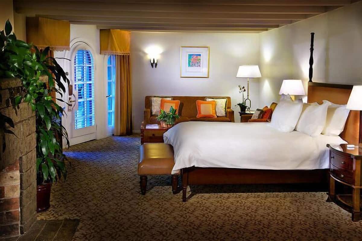 A hotel room with a large bed, bedside tables, a desk, and a chair. There is a window with curtains, a plant, and a painting on the wall.