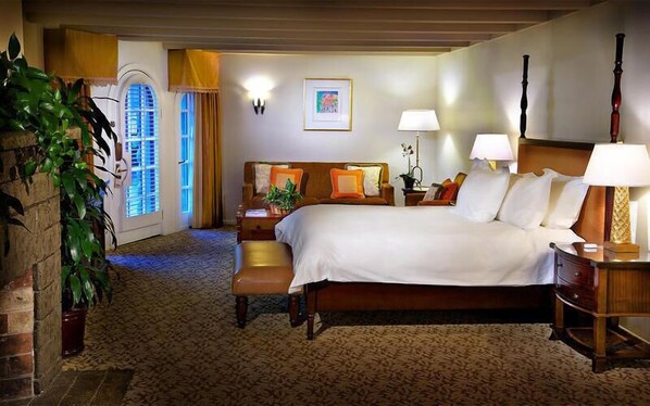 A hotel room with a large bed, bedside tables, a desk, and a chair. There is a window with curtains, a plant, and a painting on the wall.