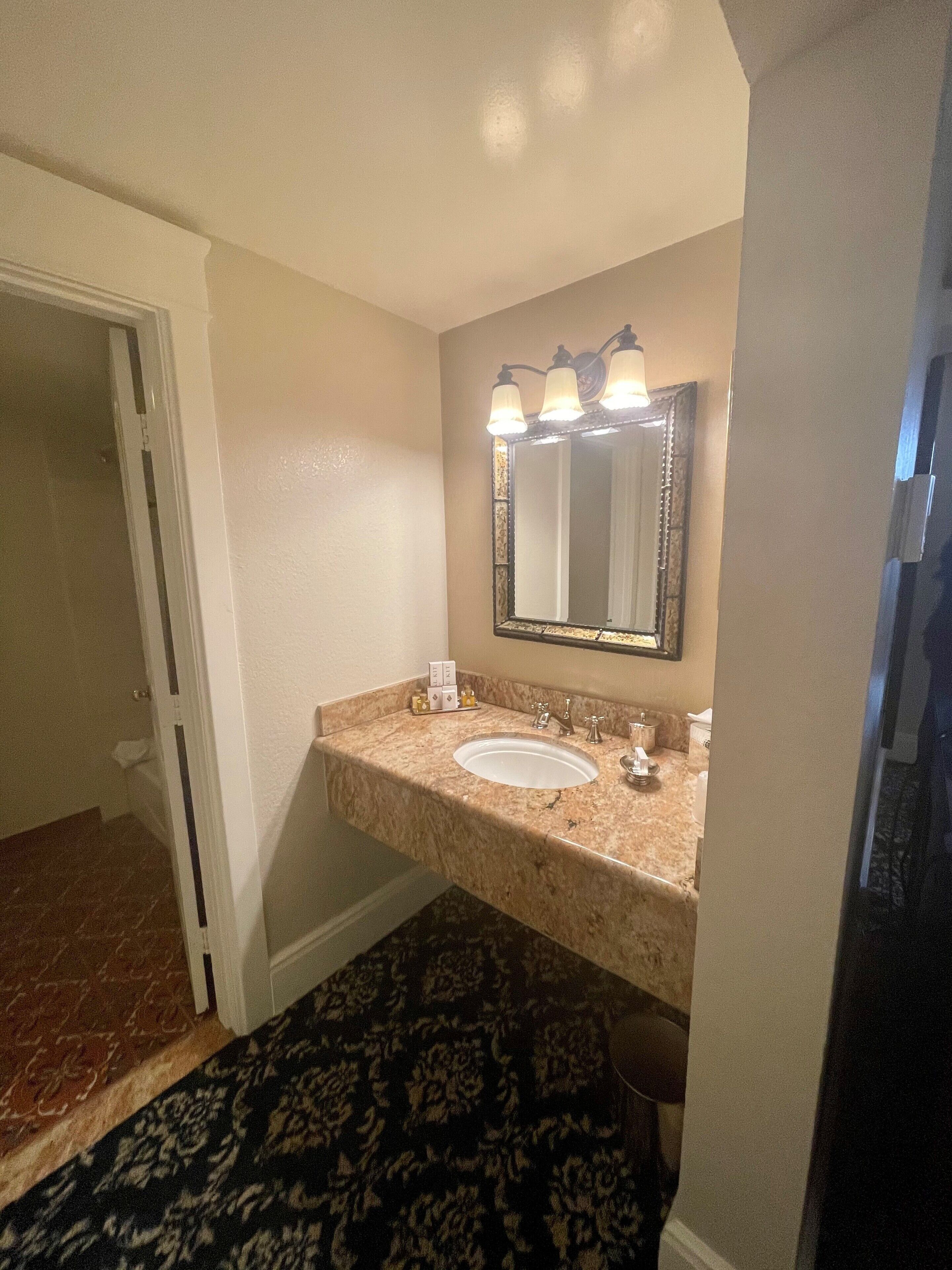 Un baño con encimera de mármol, un lavabo, un espejo con tres luces y una alfombra estampada.