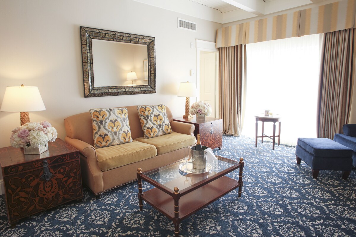 A hotel room with a beige sofa, a glass coffee table with a bottle, a side table with a lamp, and a large window with curtains.
