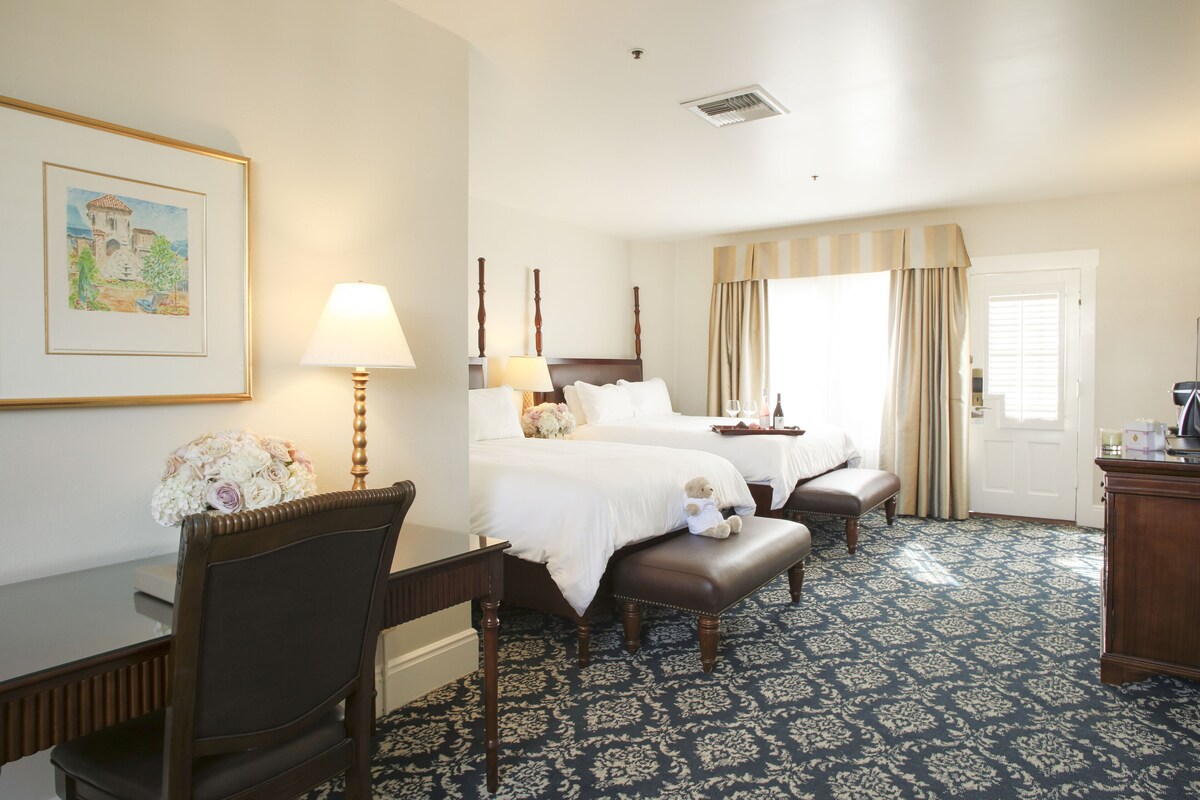 A hotel room with two beds, a desk with a lamp, a chair, and a large window with curtains.