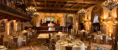 A grand banquet hall with ornate chandeliers, round tables set for dining, and a large organ in the background.