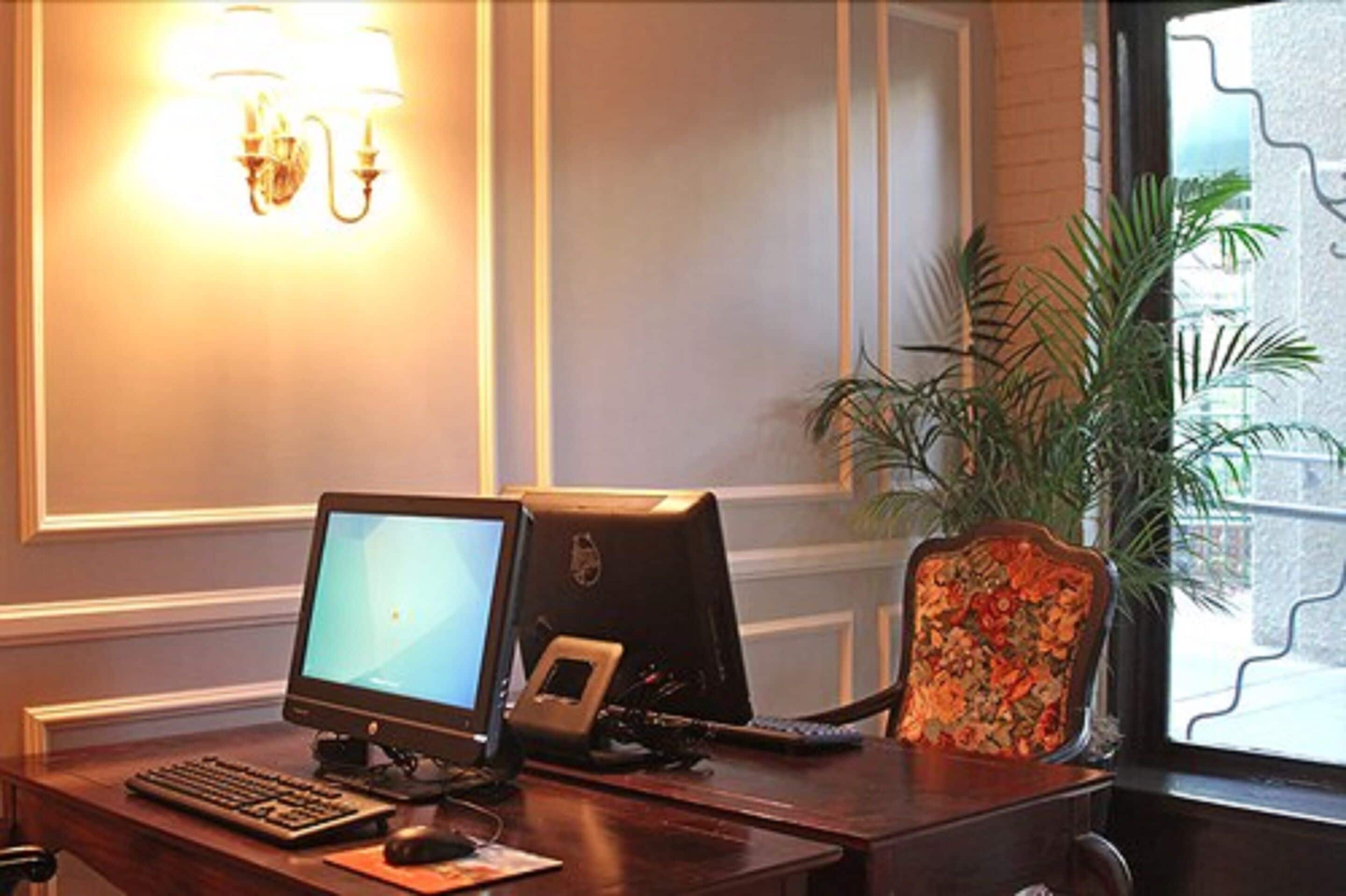 A hotel room with a desk, computer, chair, and a potted plant.