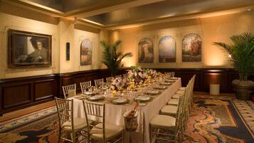 A banquet hall with a long table set for a formal event, flanked by gold chairs, and adorned with floral centerpieces.