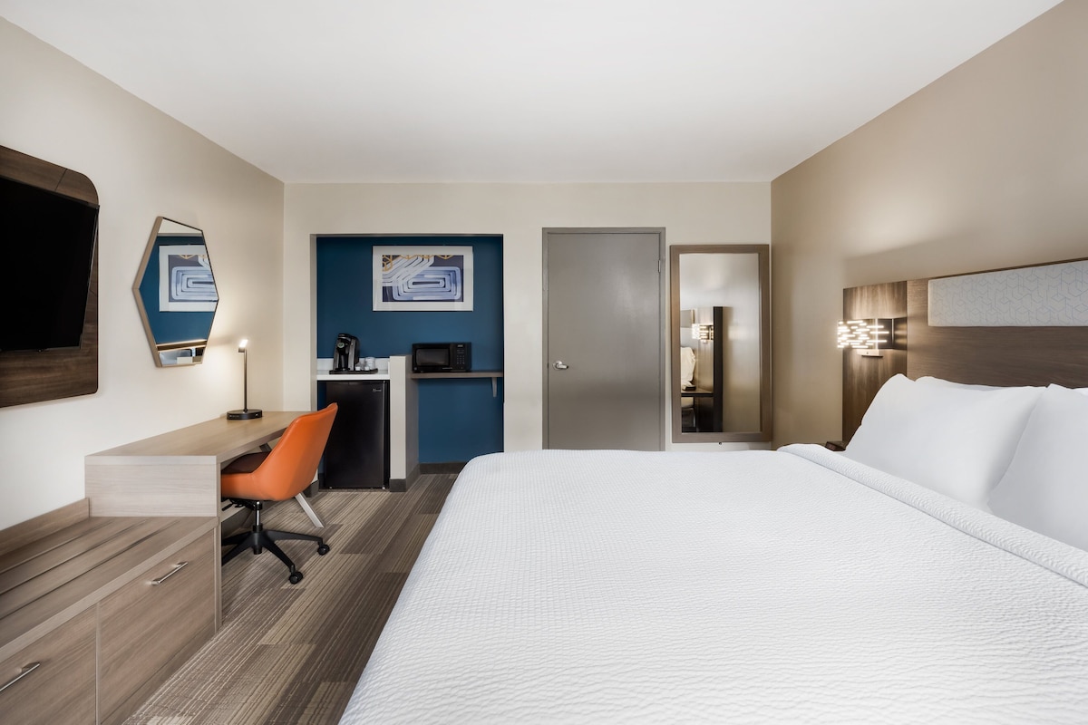 A hotel room with a bed, desk, chair, and a kitchenette area.