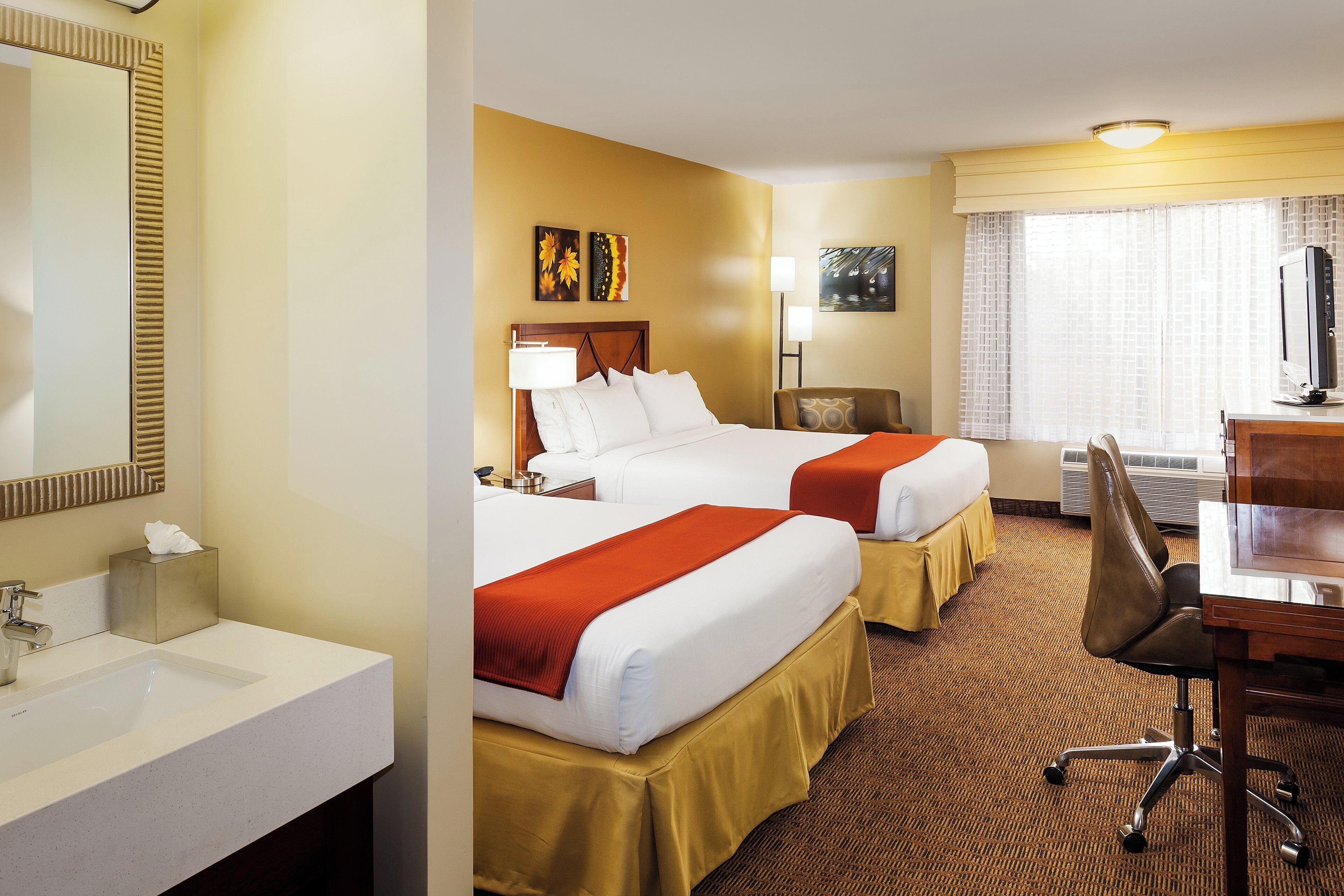 A hotel room with two beds, a desk, a chair, a TV, a lamp, and a bathroom with a sink and mirror.