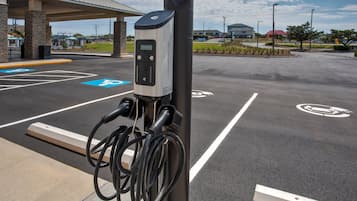 Una estación de carga con pantalla digital, conectada por cables, en un estacionamiento con espacios delimitados.