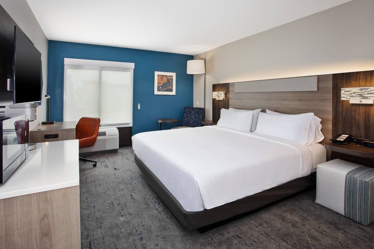 A modern hotel room with a large bed, a desk with a computer, a chair, and a TV.
