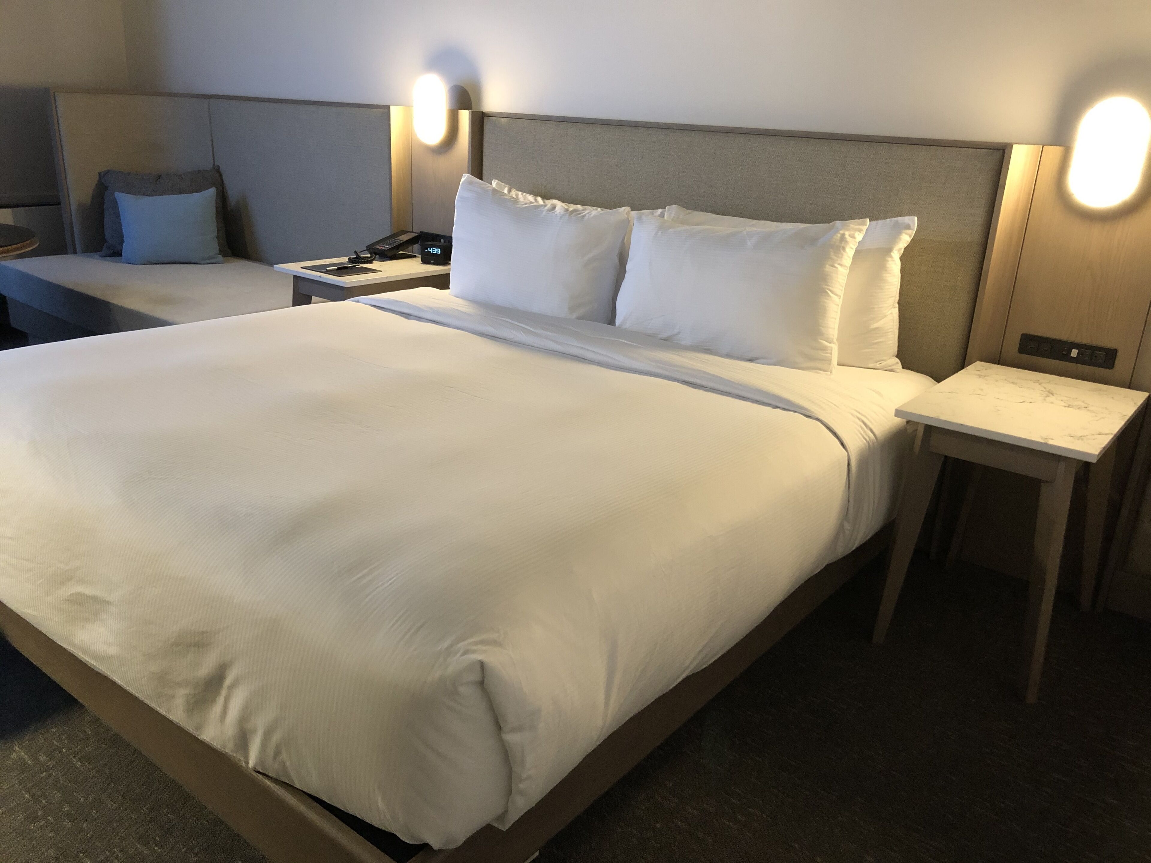 A hotel room with a large bed, two bedside tables, a nightstand, and a wall-mounted light fixture.