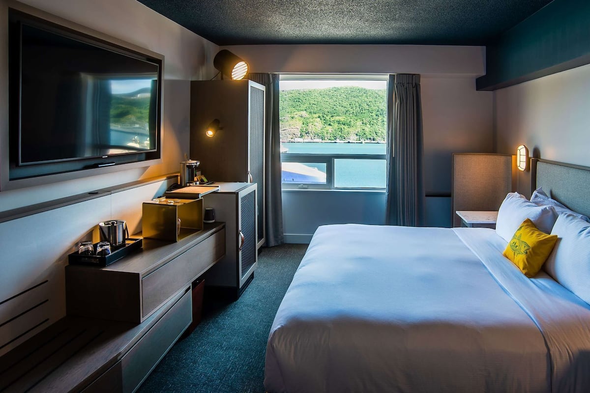 A hotel room with a bed, a TV, a desk with a coffee machine, a window with a view of greenery, and a nightstand with a lamp.