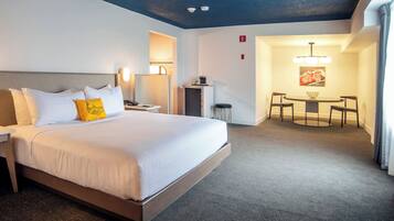 A hotel room with a large bed, bedside table, lamp, and a small kitchenette.