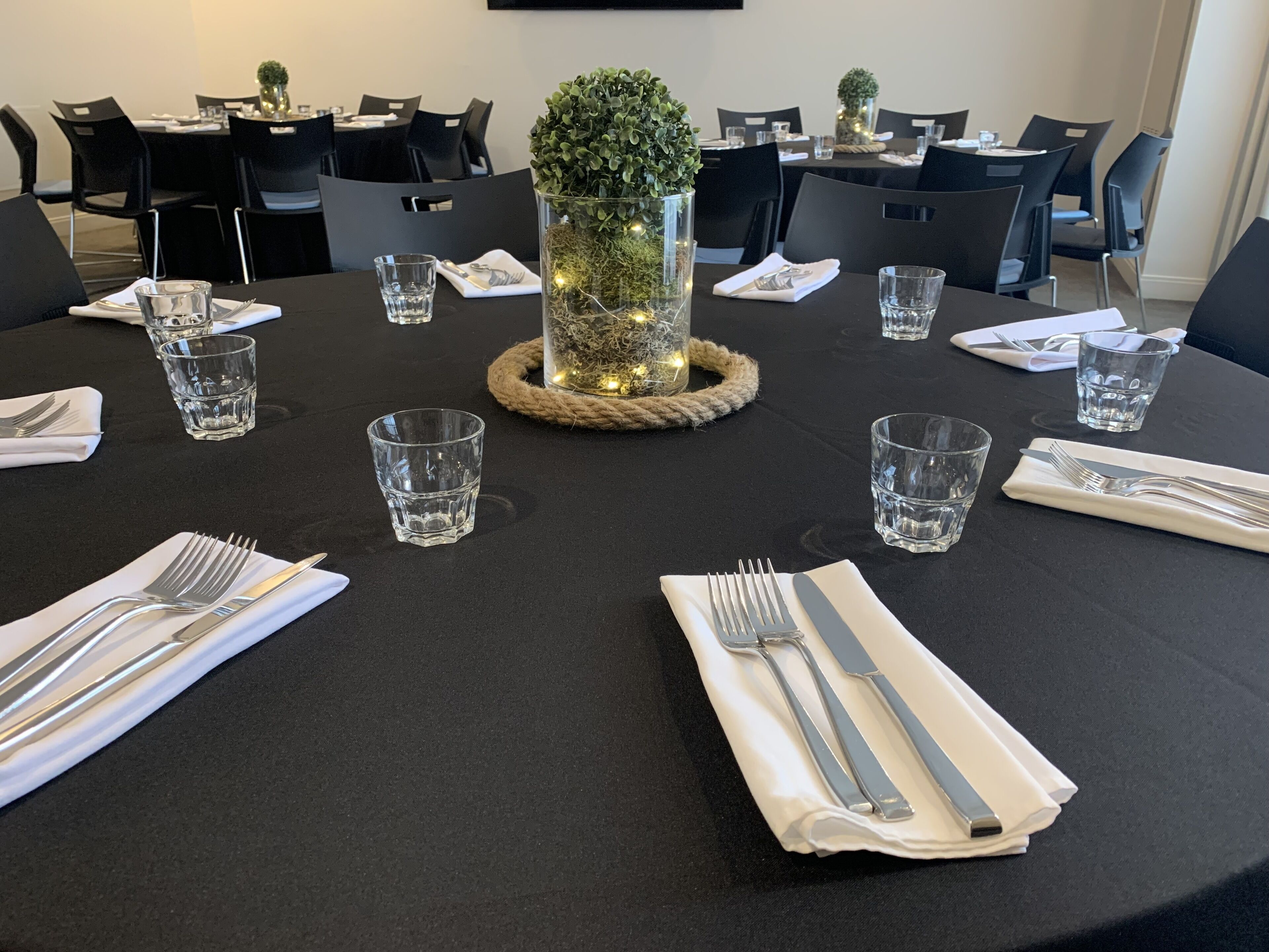 A round table set for four with clear glasses, folded napkins, and silverware. A decorative centerpiece with a small plant and lights is placed in the middle.