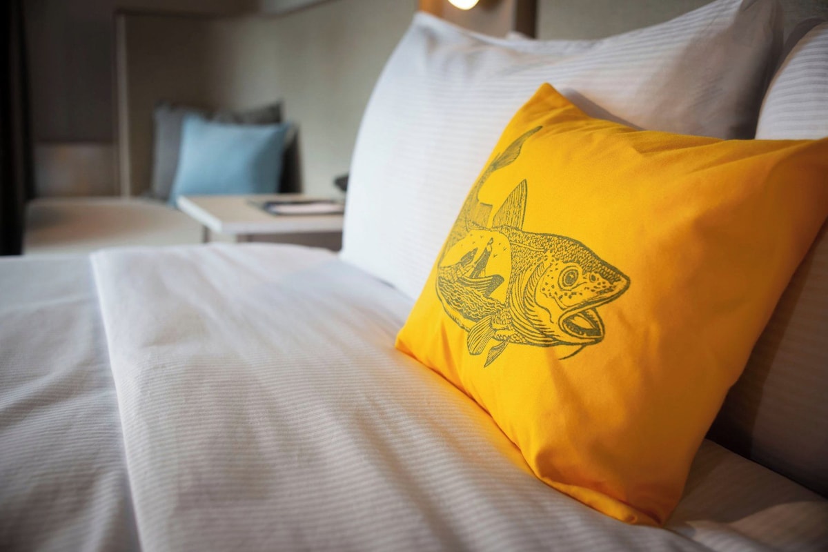 A bed with white bedding and a yellow pillow featuring a fish design.