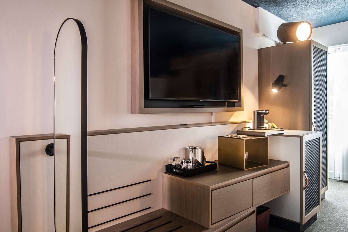 A modern hotel room with a flat-screen TV mounted above a wooden console, a glass door leading to a balcony, and a compact kitchenette.