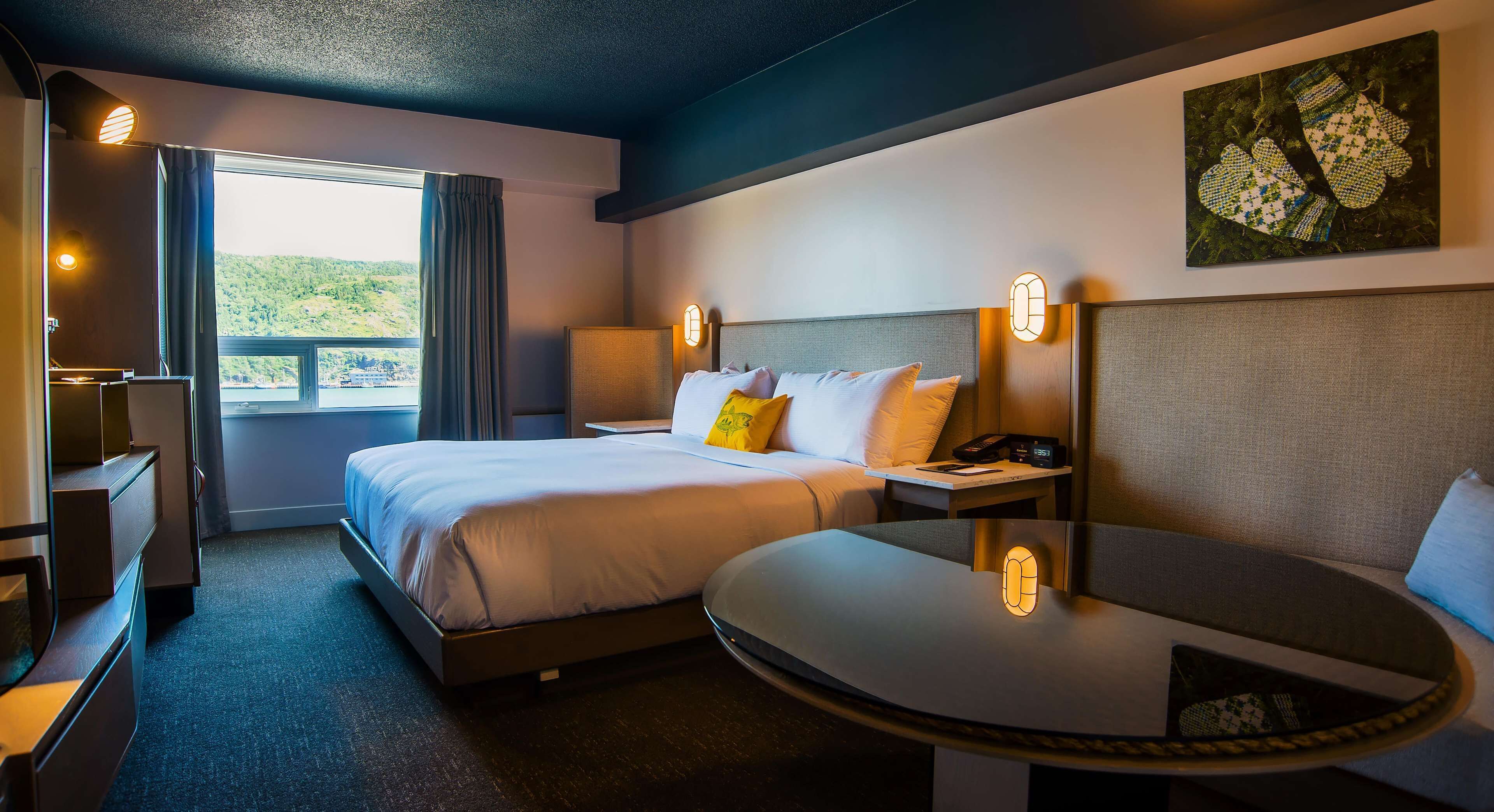 A hotel room with a bed, bedside table, lamp, and a view of the outdoors.