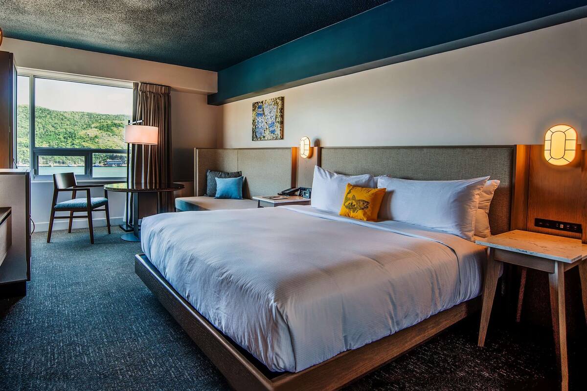 A hotel room with a large bed, a desk with a chair, a small table, and a view of the outdoors.