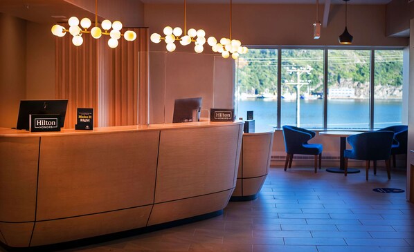 Zona de recepción de hotel con un mostrador de madera, vista a una masa de agua a través de amplios ventanales e iluminación moderna.