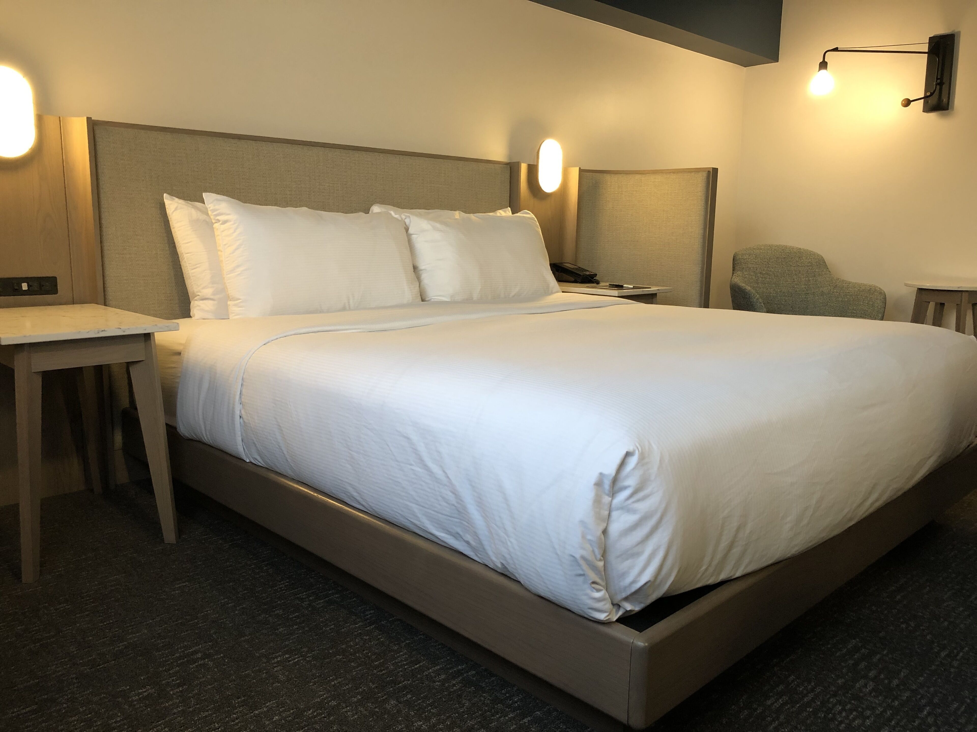 A neatly made bed with white linens, a bedside table with a lamp, and a chair in a modern hotel room.