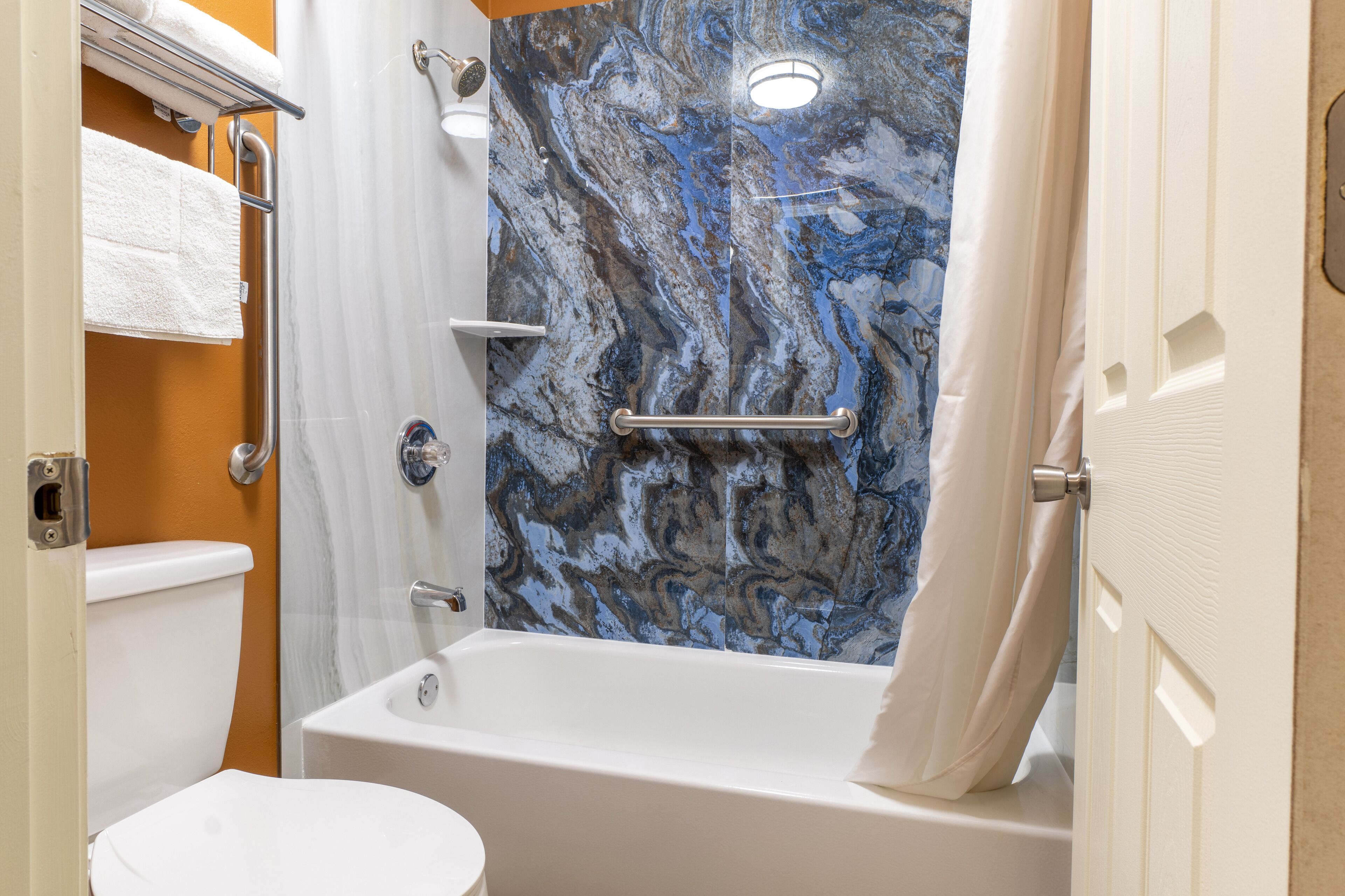 A bathroom with a marble shower wall, a white bathtub, a toilet, and a towel rack.