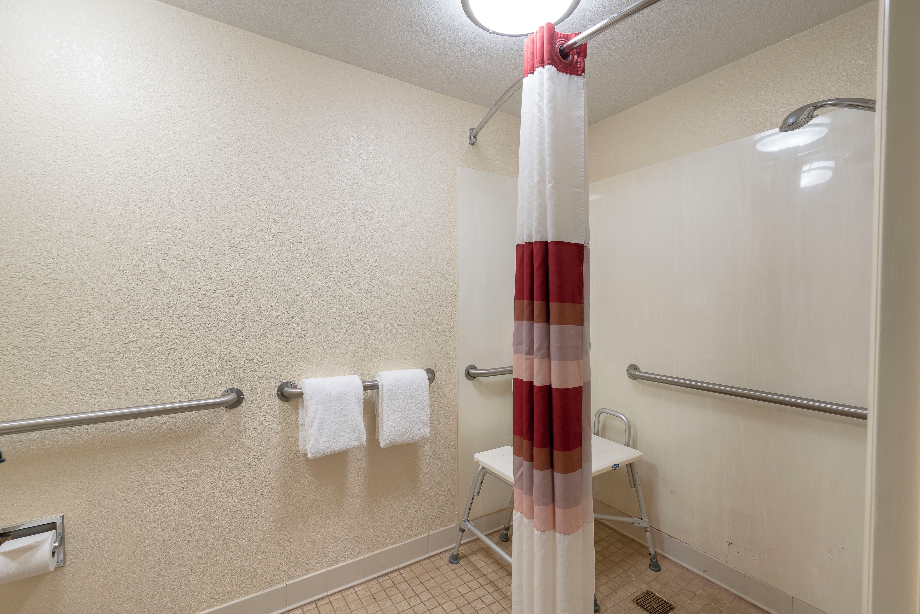 A bathroom with a shower curtain, grab bars, a towel rack with two white towels, and a wheelchair-accessible shower seat.