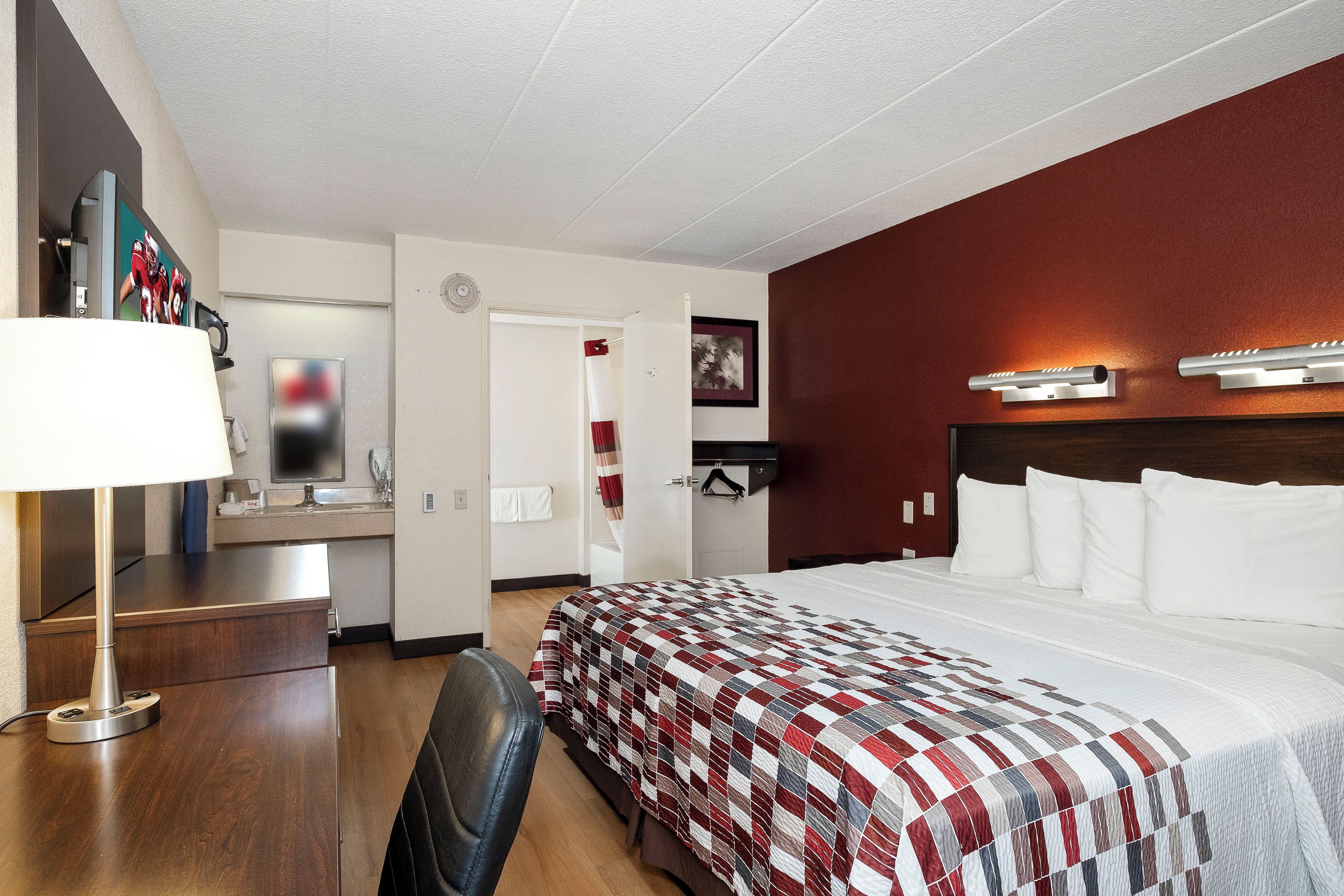 A hotel room with a bed, a desk, a chair, a TV, and a bathroom visible through an open door.