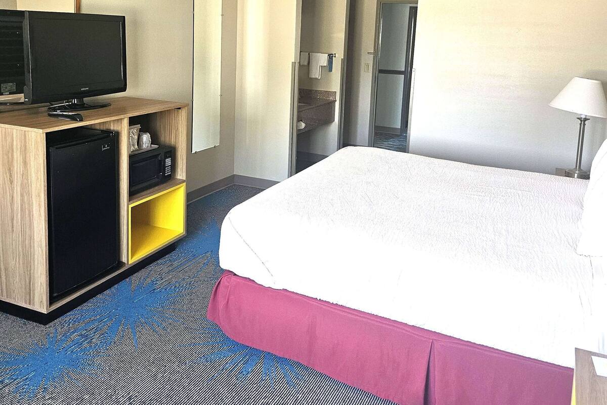 Habitación de hotel con cama, televisor sobre un soporte de madera, un refrigerador pequeño y un baño visible a través de una puerta abierta.
