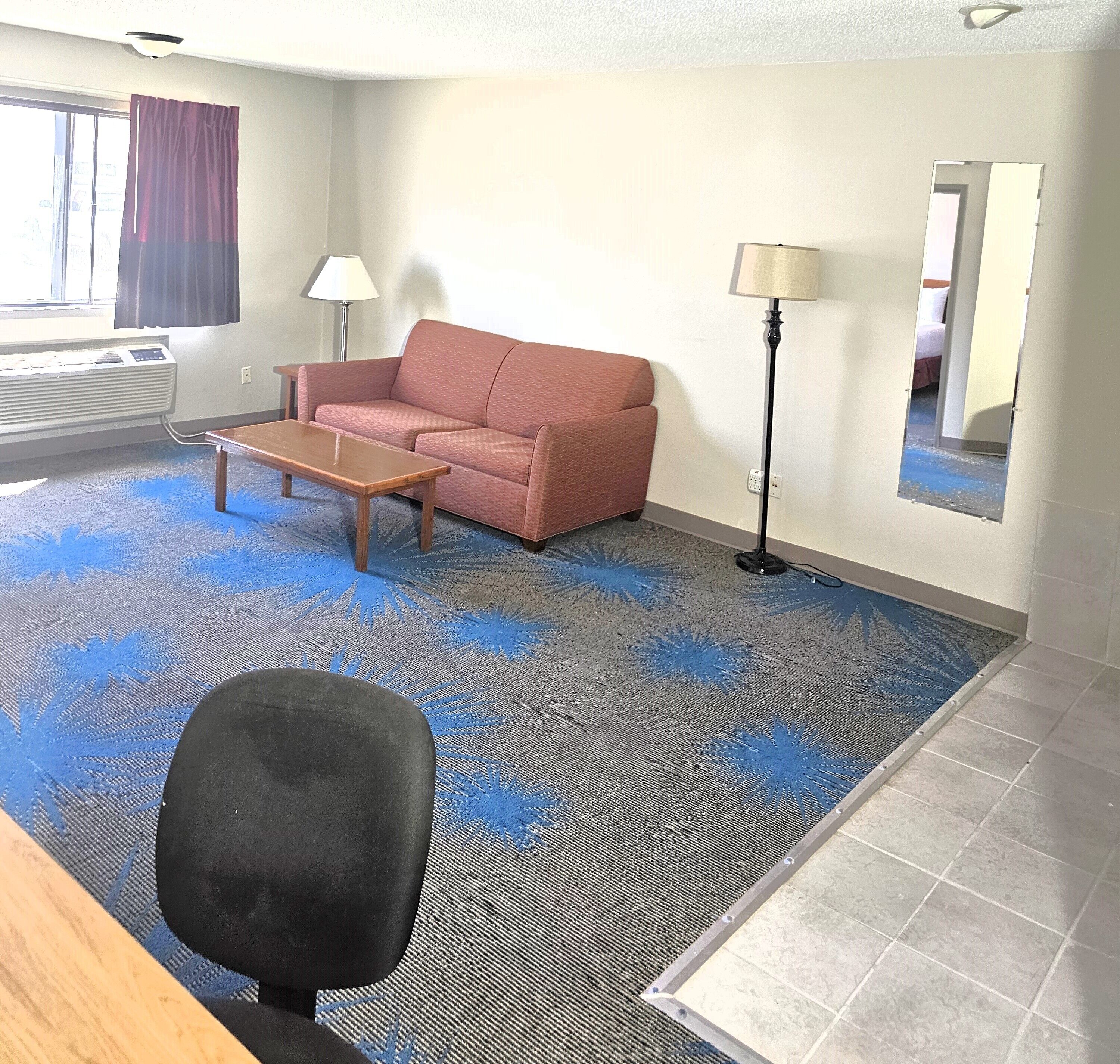 Habitación de hotel con un sofá rojo, una mesa de centro de madera, una silla de oficina negra, una lámpara de pie y un espejo grande en la pared.