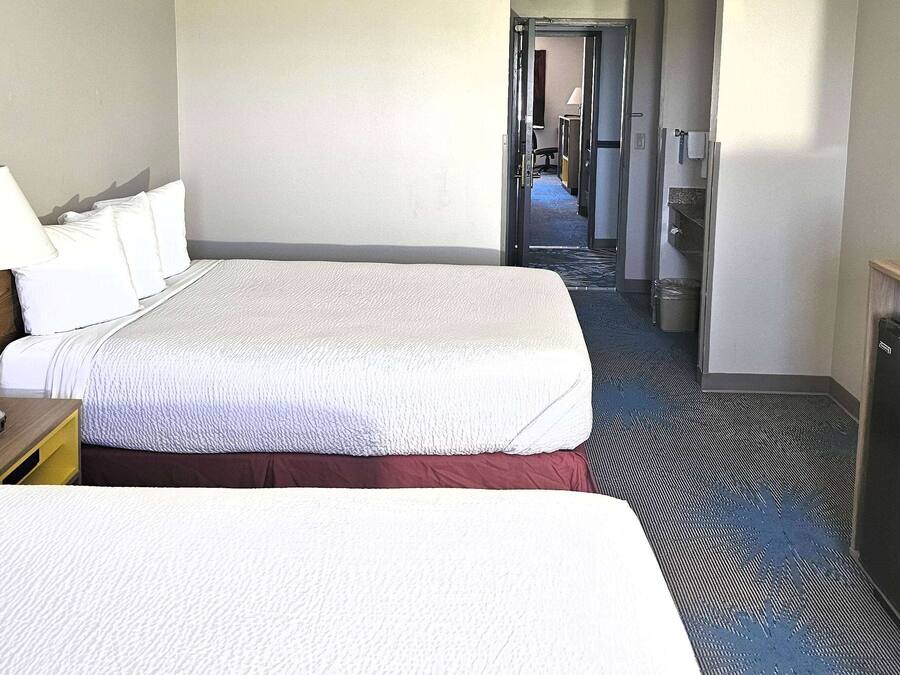 A hotel room with two beds, a desk with a lamp, and a hallway leading to other rooms.