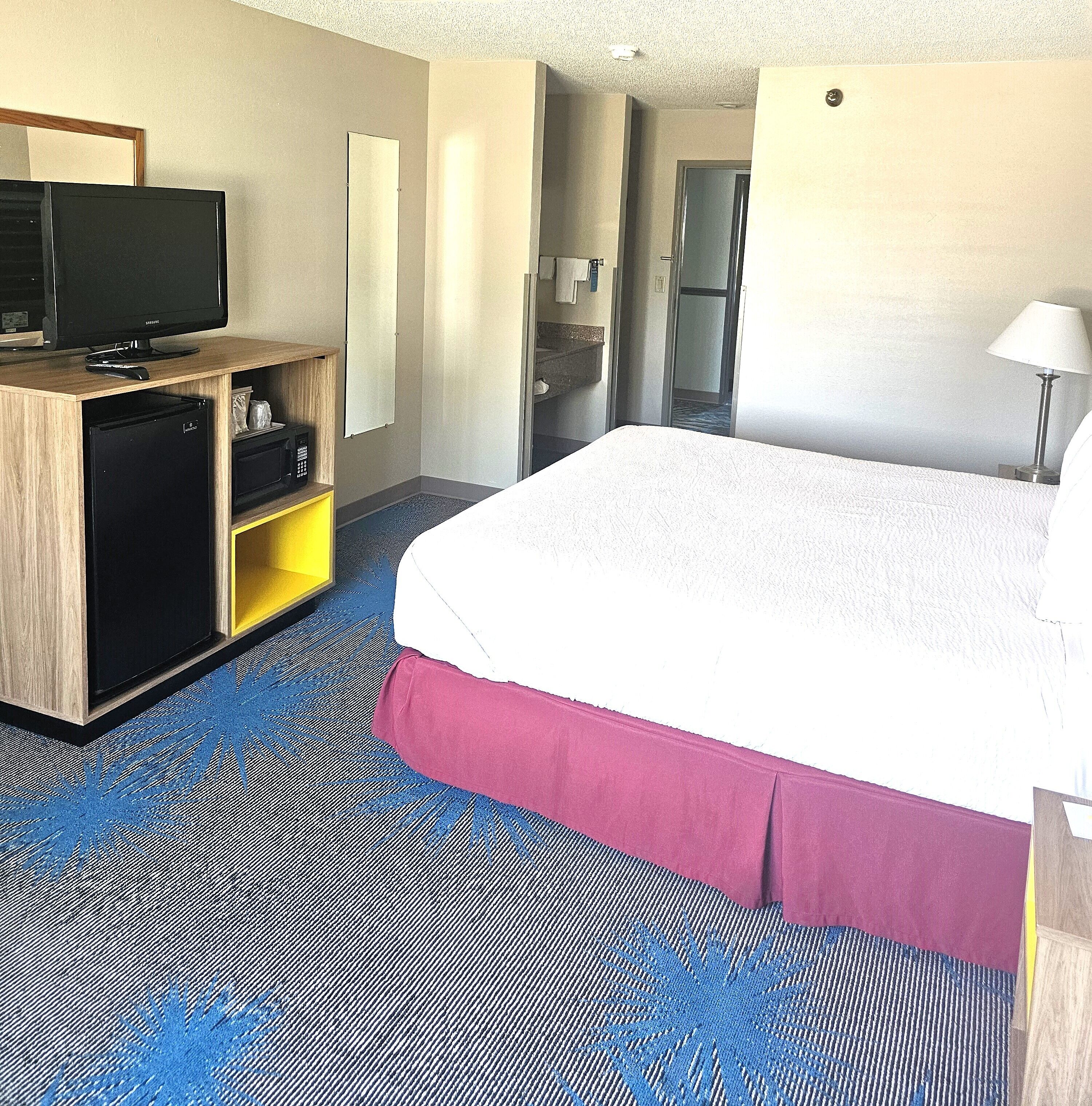 A hotel room with a bed, a desk with a television, a minibar, and a bathroom visible through an open door.