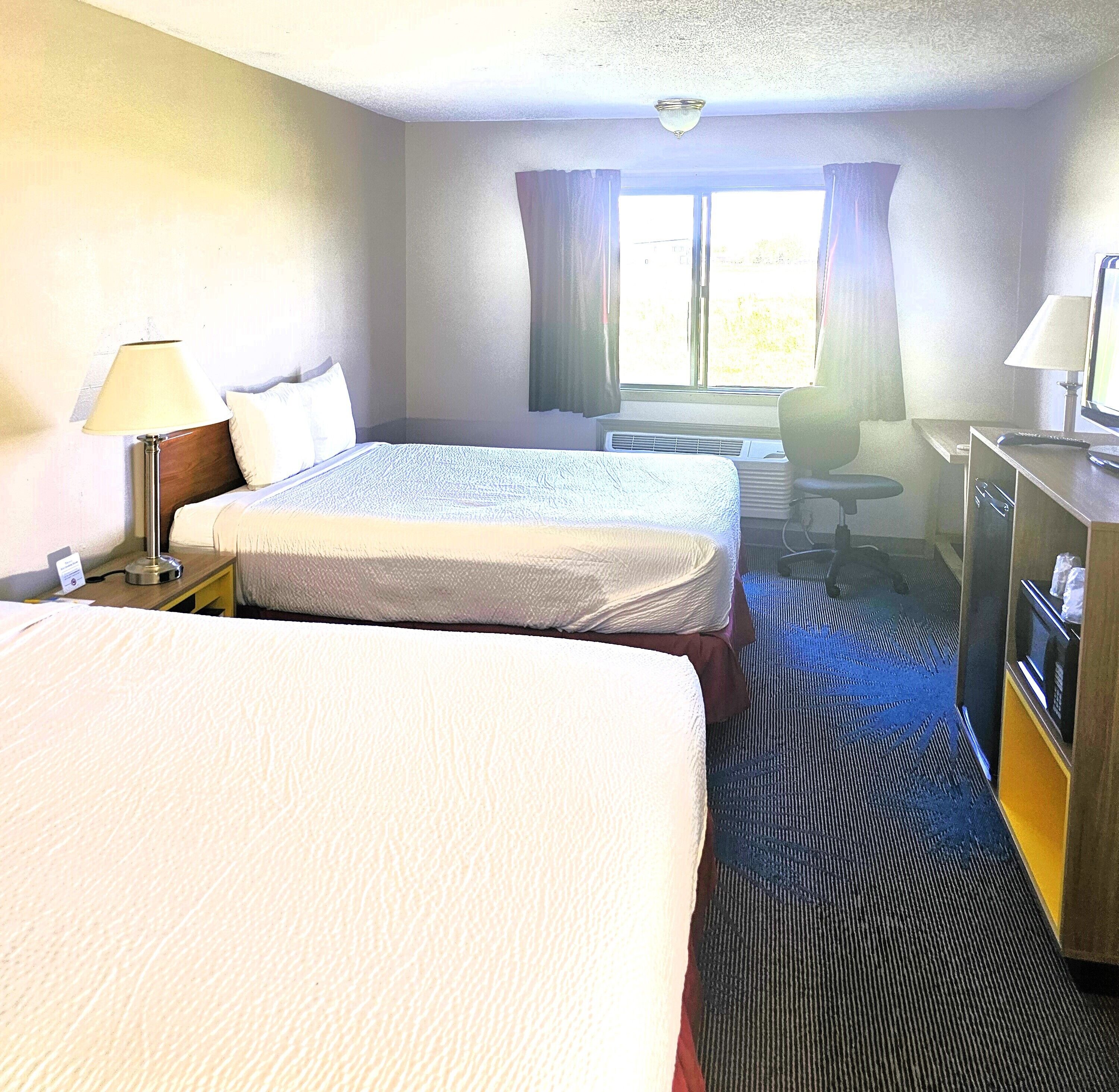 A hotel room with two beds, a desk, a chair, a lamp, and a window with curtains.