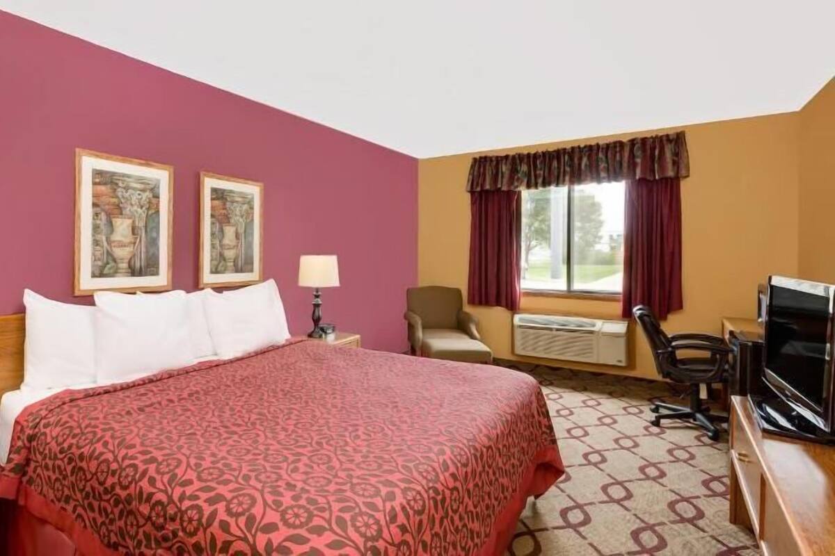 Habitación de hotel con cama, dos lámparas de noche, una silla, un escritorio con computadora y dos cuadros en la pared.