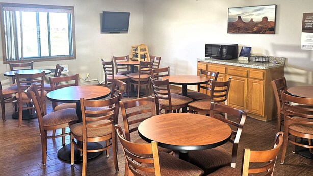A dining area with multiple wooden tables and chairs, a kitchen area with a microwave and a cabinet, and a large window with curtains.