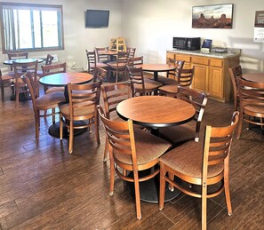 A dining area with multiple wooden tables and chairs, a kitchen area with a microwave and a cabinet, and a large window with curtains.