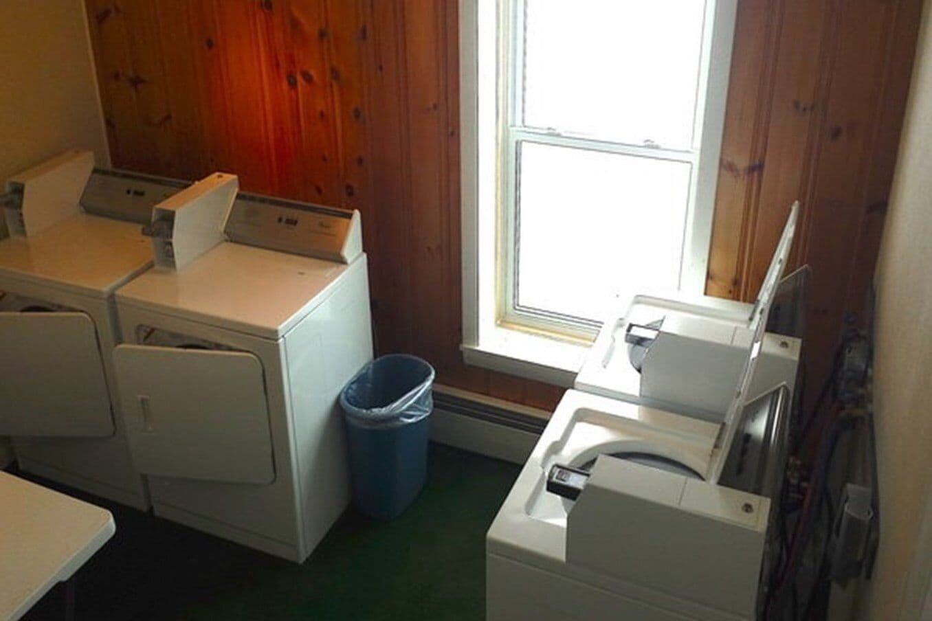 Une buanderie équipée de trois machines à laver, d’une poubelle bleue et d’une fenêtre dont les murs sont recouverts de panneaux en bois.