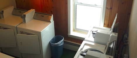 A laundry room with three washing machines, a blue trash bin, and a window with wooden paneled walls.