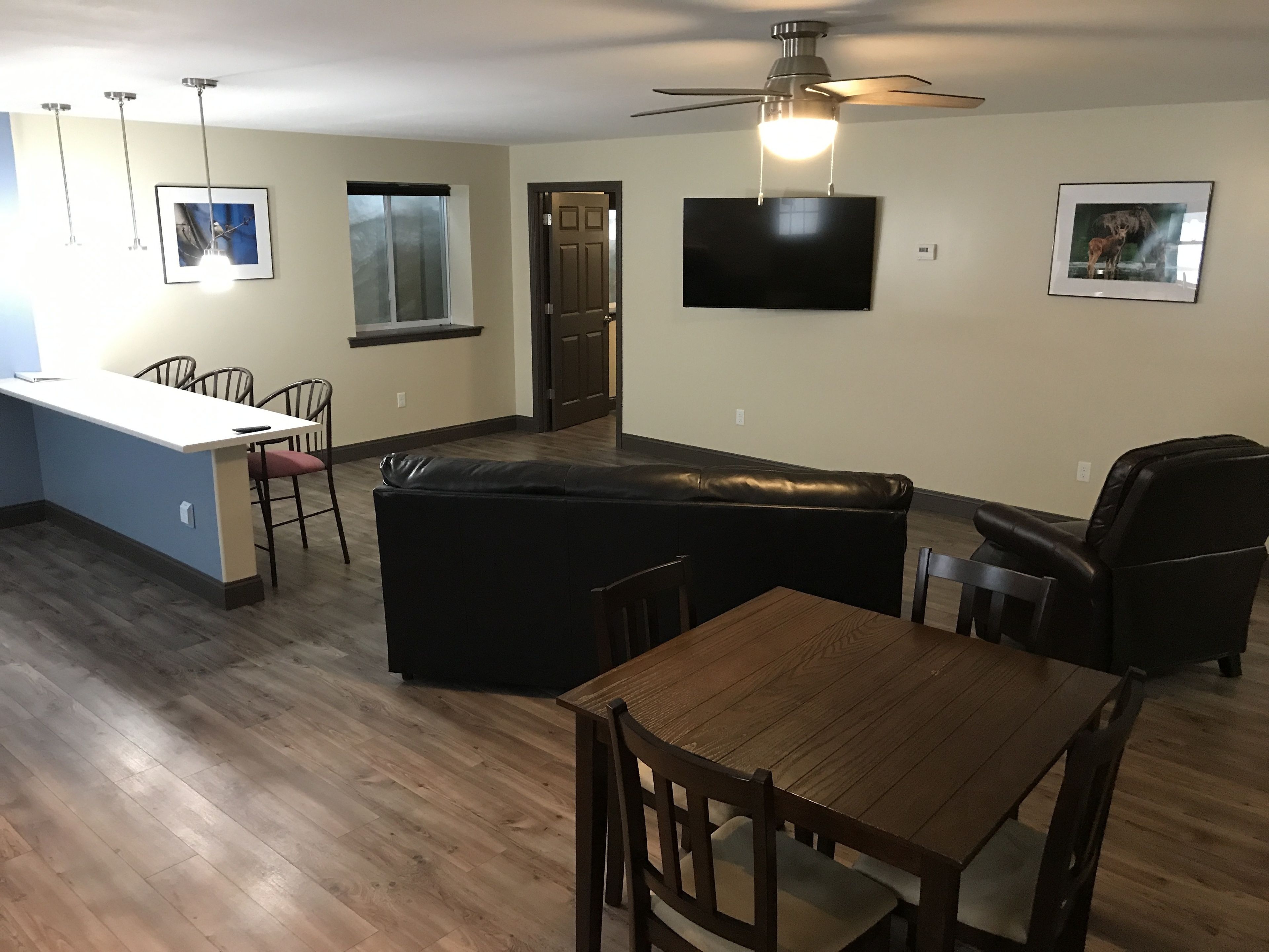 A modern living room with a black leather sofa, wooden dining table, and chairs. A flat-screen TV is mounted on the wall, and there are framed pictures on the walls.