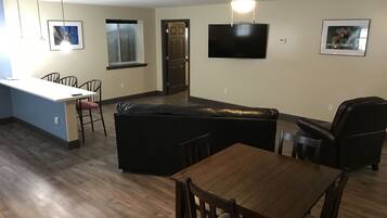 Una sala de estar moderna con un sofá de cuero negro, una mesa de comedor de madera y sillas. Un televisor de pantalla plana está montado en la pared, y hay cuadros enmarcados adornando las paredes.