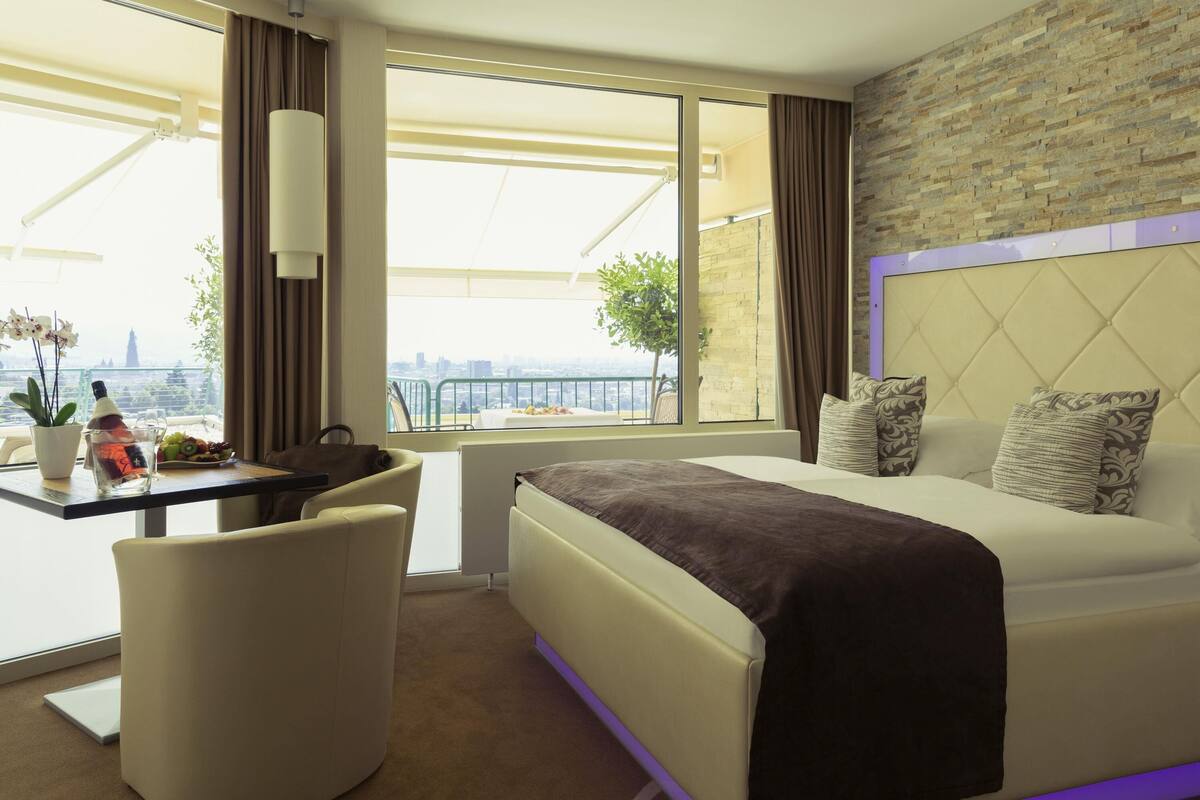 Ein Hotelzimmer mit Bett, Schreibtisch mit Stuhl, einer Topfpflanze und Blick auf die Stadt.