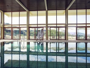 A large indoor swimming pool with clear glass walls, a wooden ceiling, and a view of outdoor seating and greenery. - Mercure Hotel Panorama Freiburg (Freiburg im Breisgau)