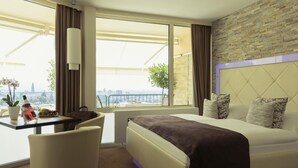 Ein Hotelzimmer mit Bett, Schreibtisch mit Stuhl, einer Topfpflanze und Blick auf die Stadt.