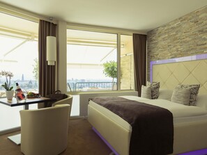 Hotelkamer met een bed, een bureau met stoel, een potplant en uitzicht op de stad.