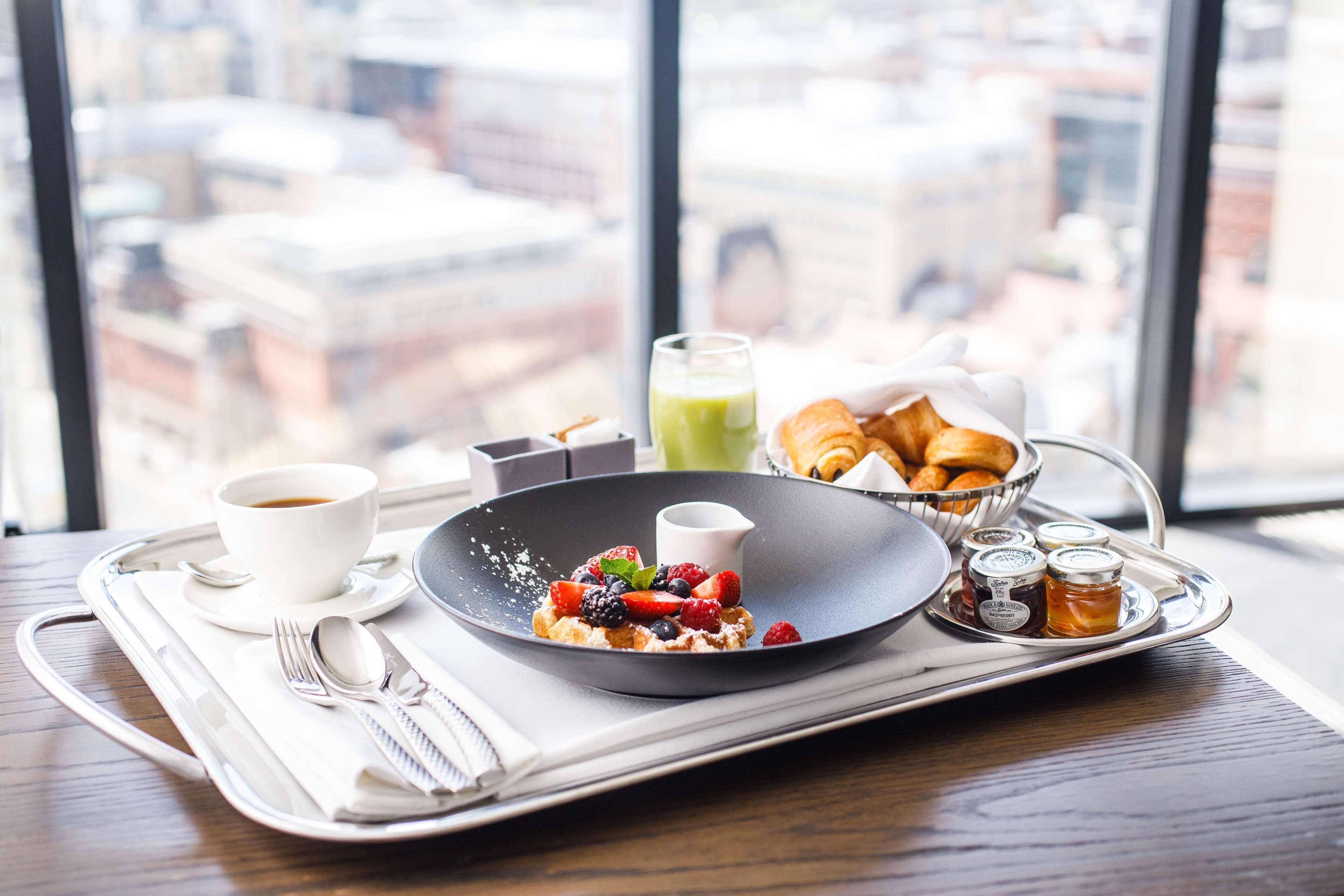 A breakfast tray with a plate of pancakes topped with berries, a cup of coffee, a glass of juice, croissants, and condiments on a tray.