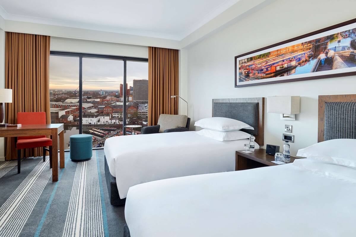A hotel room with two beds, a desk, a chair, a lamp, and a large window with a city view.