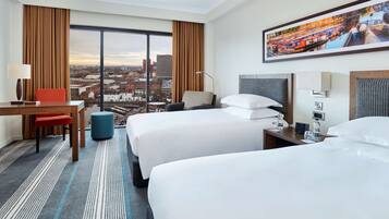 Habitación de hotel con dos camas, un escritorio, una silla, una lámpara y un ventanal con vistas a la ciudad.