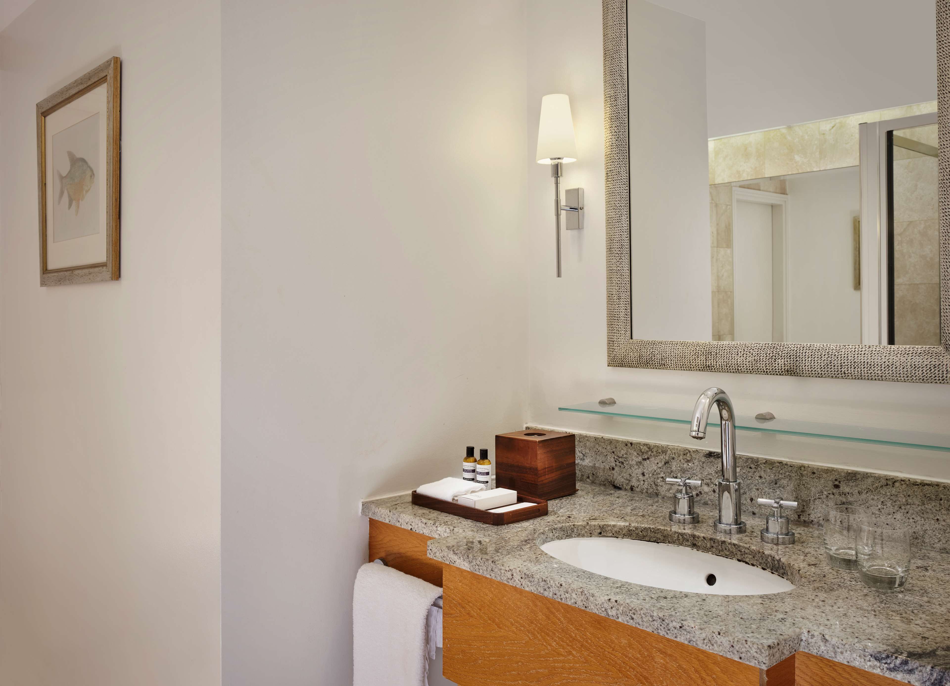 Un baño con encimera de granito, un lavabo blanco, un espejo con marco texturizado, una lámpara de pared e imagen enmarcada.